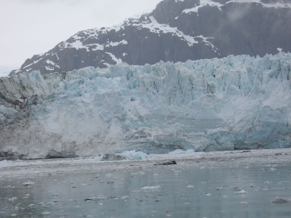 Massive glacier face with ice chunks in foreground water, snow-capped mountain in background