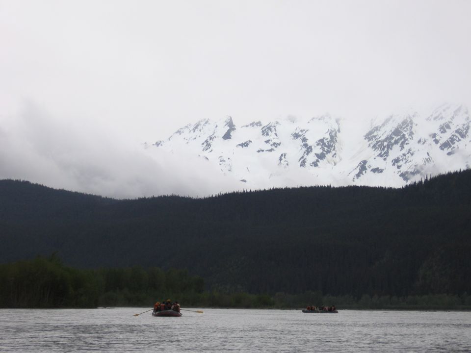 Two rafts with people paddling on a wide river, dark forest on the shore, and snow-capped mountains in the background under a cloudy sky