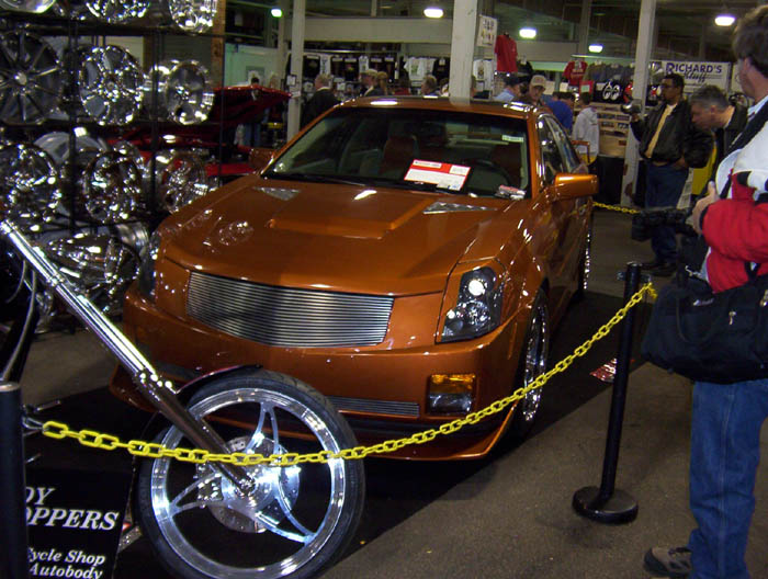 Copper custom sedan with billet grille at indoor car show, chrome chopper wheel in foreground