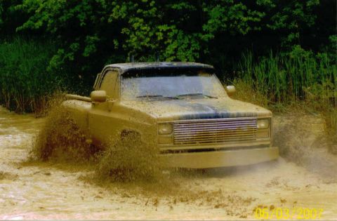 1976 Chevrolet Blazer - Muddy pickup truck navigates through water, kicking up spray. The grille suggests a 1975 model year.