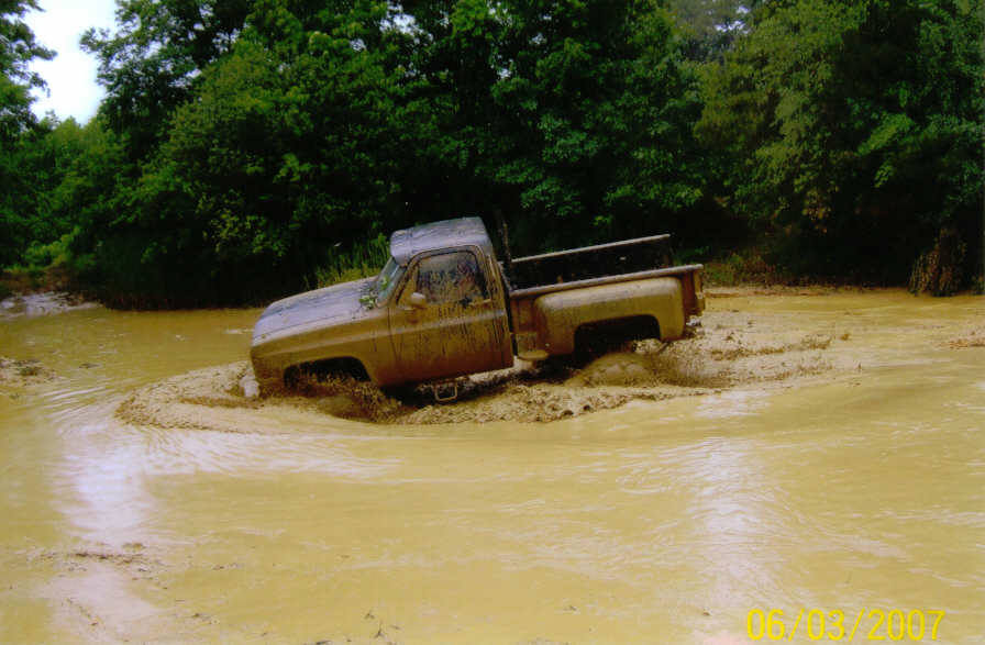 Muddy pickup truck driving through a shallow body of water, trees in background