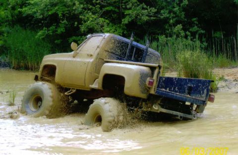 1976 Chevrolet Blazer - A lifted pickup truck navigates a muddy water obstacle with large tires churning up the water.