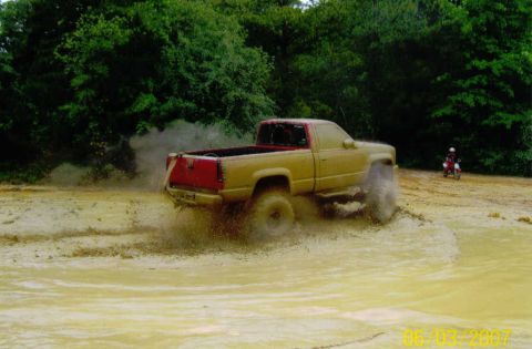 1976 Chevrolet Blazer - Pickup truck navigating a muddy off-road course, with a dirt bike rider in the distance.