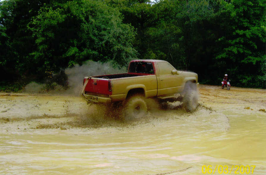 Pickup truck splashing through a muddy puddle with trees in the background. A person on a dirt bike is visible to the right