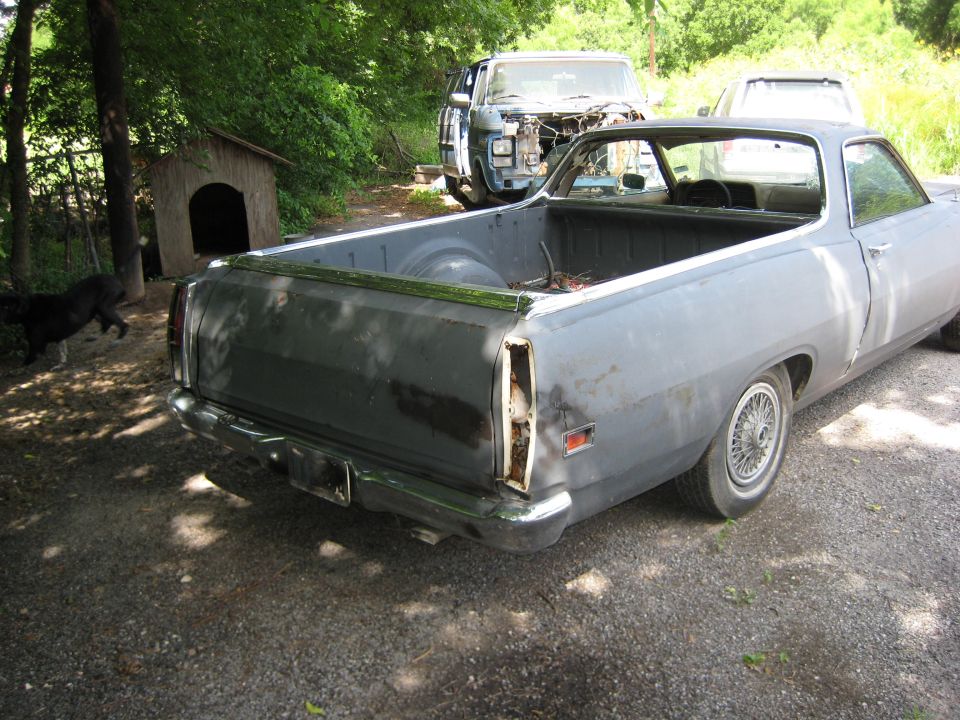 Rear 3/4 view of a gray primer Ford Ranchero with rusty tailgate on gravel, doghouse and old truck behind