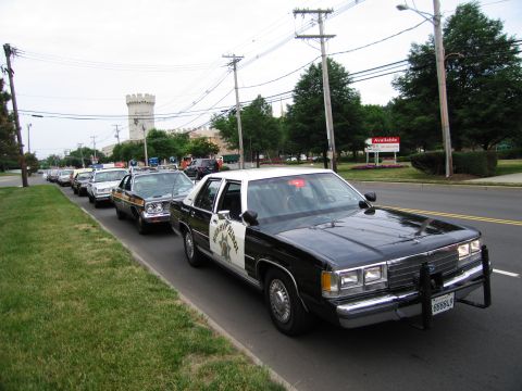 1991 Ford LTD Crown Victoria - black-and-white highway patrol sedan with a star emblem leads a roadside line of vintage police cars near a castle-like building.