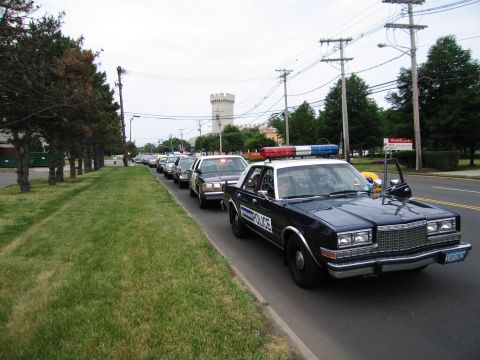 1991 Ford LTD Crown Victoria - dark blue POLICE sedan with a roof light bar leads a procession of classic police cars along a grass-lined roadway.