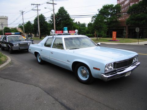 1991 Ford LTD Crown Victoria - light-blue 1970s STATE POLICE four-door sedan with roof beacons drives down a road ahead of a black patrol car.