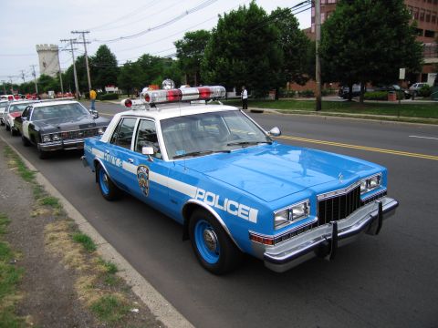 1991 Ford LTD Crown Victoria - bright blue and white POLICE sedan with a gold shield and roof light bar parked at a curb, leading a line of classic patrol cars.