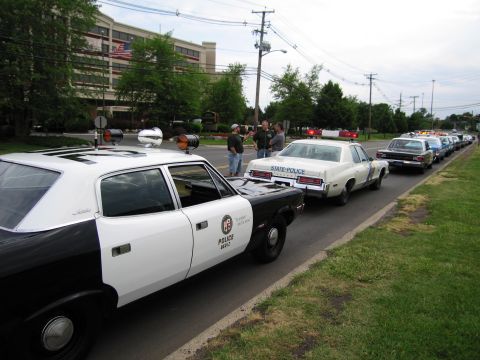 1991 Ford LTD Crown Victoria - black-and-white police car and a white STATE POLICE sedan parked along a roadside line of vintage patrol cars, with people standing nearby.