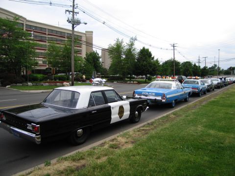 1991 Ford LTD Crown Victoria - black-and-white classic police sedan leads a long line of vintage cruisers, including a blue NYPD car, parked along a roadside.