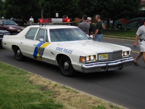 1991 Ford LTD Crown Victoria - white mid-1970s State Police sedan marked with blue and yellow striping and a roof lightbar is parked at the curb as people walk past.