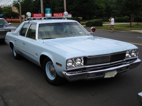 1991 Ford LTD Crown Victoria - light-blue 1970s Plymouth State Police sedan with a roof lightbar sits at the curb in front of a State Police banner.