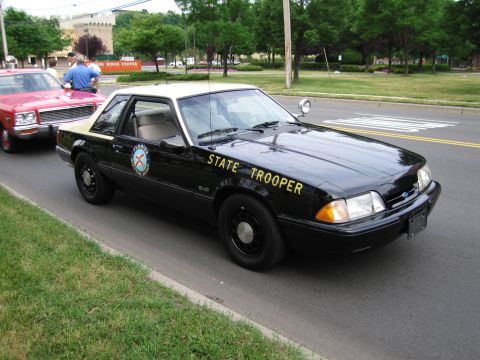 1991 Ford LTD Crown Victoria - black fox-body Ford Mustang 5.0 marked State Trooper with gold lettering sits at the curb next to a red classic car.