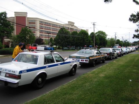 1991 Ford LTD Crown Victoria - yellow vintage police sedan with a blue stripe and roof lightbar leads a long line of classic cruisers parked along a roadside.