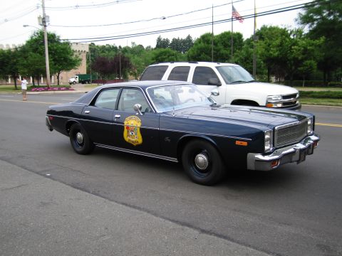 1991 Ford LTD Crown Victoria - dark-blue 1970s police sedan bearing a gold door shield is parked at the curb beside a white pickup, an American flag flying behind.