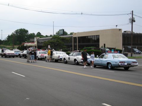 1991 Ford LTD Crown Victoria - long row of classic police cruisers is parked along a street in front of a low office building as people stand beside them.