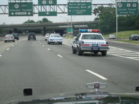 1991 Ford LTD Crown Victoria - North Haven Police sedan leads other cruisers along a highway beneath green overhead signs for the Lincoln Tunnel and Jersey City.