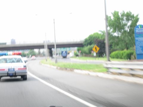 1991 Ford LTD Crown Victoria - motion-blurred view from inside a moving car shows a white police cruiser ahead on a curving highway with a merge sign.