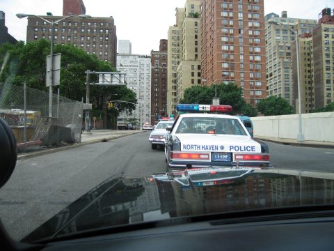 1991 Ford LTD Crown Victoria - North Haven Police cruiser with lightbar drives down a city street flanked by tall apartment buildings, seen over a car hood.