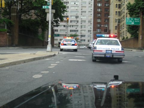 1991 Ford LTD Crown Victoria - North Haven Police cruiser with flashing lights turns at a city intersection near tall apartment buildings and street signs.