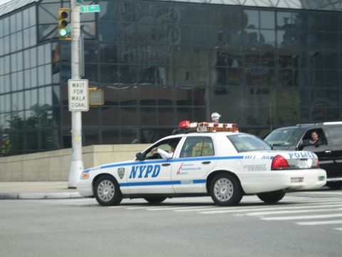1991 Ford LTD Crown Victoria - white NYPD Ford Crown Victoria patrol car with blue striping crosses a city intersection in front of a glass building.