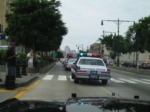 1991 Ford LTD Crown Victoria - North Haven Police cruiser with lightbar drives down a tree-lined city avenue as pedestrians wait with luggage on the sidewalk.