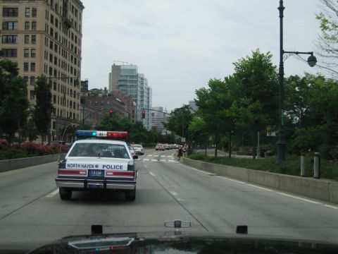 1991 Ford LTD Crown Victoria - North Haven Police cruiser leads traffic down a divided city boulevard lined with trees and tall buildings.