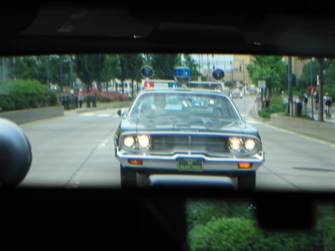 1991 Ford LTD Crown Victoria - Seen through a windshield, a dark classic police car with headlights and roof lights on follows on a tree-lined city street.