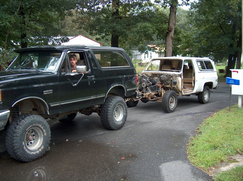 Lifted green Ford Bronco towing a stripped hoodless white SUV on a tow bar