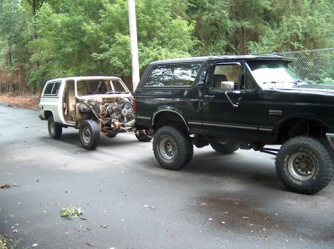 1988 Ford Bronco - Dark green full-size Ford Bronco on a suspension lift with mud-terrain tires on white and black steel wheels, hooked to a tow bar pulling a…