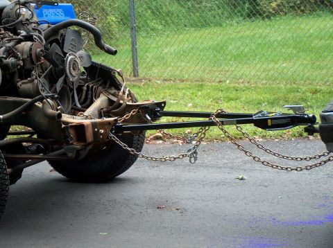 1988 Ford Bronco - A stripped, body-less truck chassis with its V8 engine, fan clutch and radiator hoses still in place is hitched to a black A-frame tow bar…