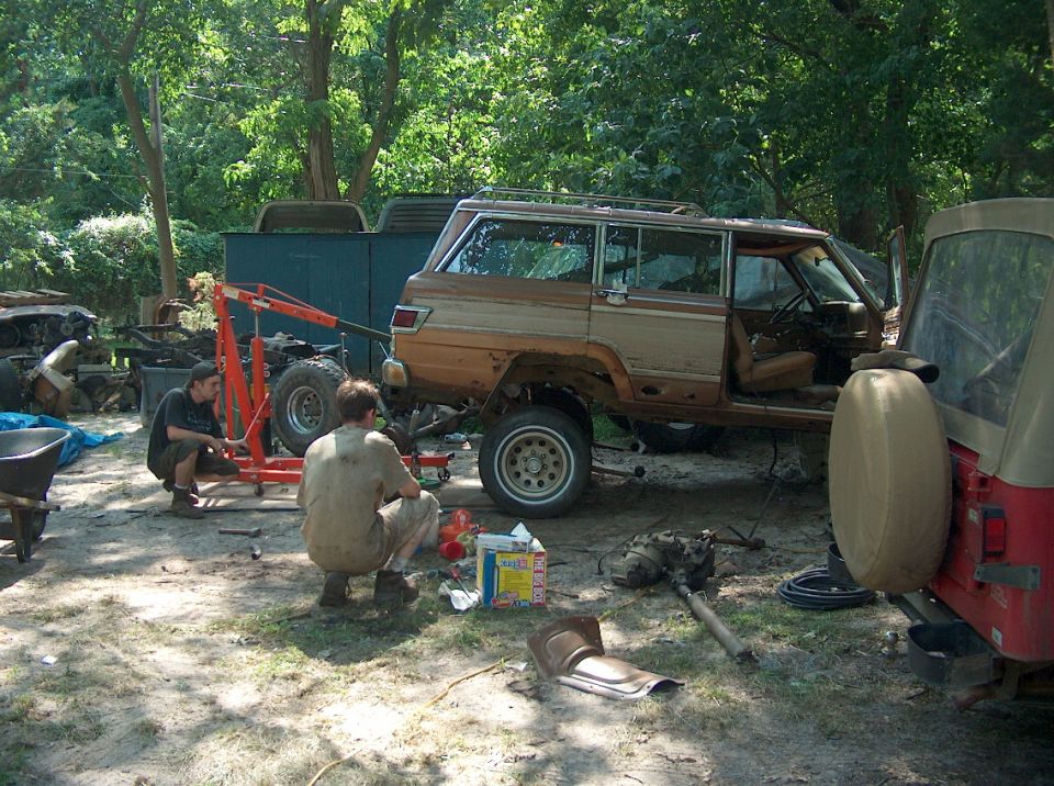Two guys working on a wood-paneled Jeep Grand Wagoneer with an engine hoist nearby. Another Jeep is parked to the right