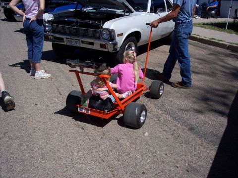 1972 Ford Ranchero - A man pulls a wagon with two children past a white car at an outdoor event.