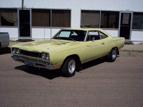 1972 Ford Ranchero - A pale yellow Plymouth Road Runner is parked in front of a white building with large windows.
