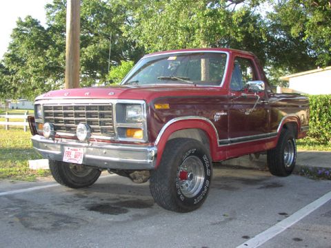 1981 Ford Bronco - This red Ford pickup truck is parked in a lot with trees and a building in the background.