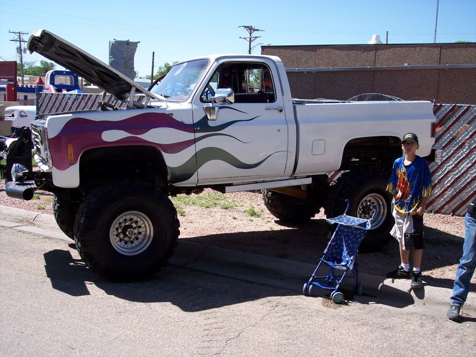 Lifted white pickup truck with purple and green flame graphics, hood open. A boy stands next to it