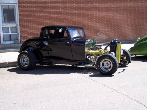 1972 Ford Ranchero - A black hot rod coupe with a visible engine sits parked on a street next to a green vehicle.