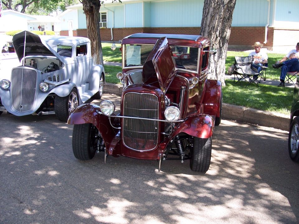Maroon hot rod coupe with hood open, parked next to a gray hot rod truck. People sit in chairs in the background