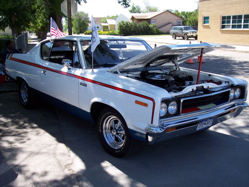 White 1970 AMC Rebel Machine coupe with red and blue side stripes, hood open, parked on a street