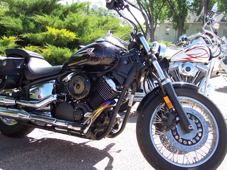 Black motorcycle front view with chrome exhaust and wire wheels. Another motorcycle is visible behind it