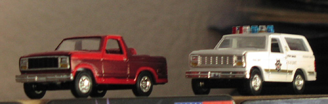 Two toy trucks on a shelf: a red pickup and a white police SUV with lights. The police SUV has text and a badge