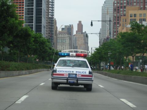 1991 Ford LTD Crown Victoria - North Haven Police cruiser with lightbar drives down a wide road toward a dense city skyline of high-rise buildings.