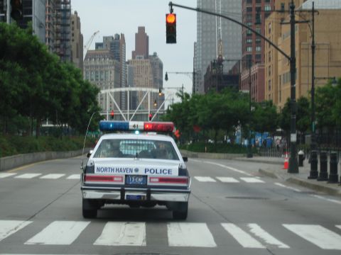1991 Ford LTD Crown Victoria - North Haven Police cruiser sits at a crosswalk under a red traffic light amid downtown high-rises and a domed structure.