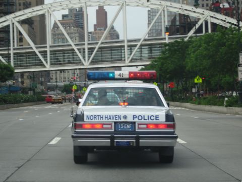 1991 Ford LTD Crown Victoria - white and blue North Haven Police sedan with a roof lightbar drives down a city avenue beneath a white arched truss bridge.