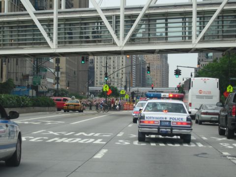 1991 Ford LTD Crown Victoria - North Haven Police sedan with flashing lights drives through a downtown intersection near a yellow taxi and a bus beneath a glass skywalk.