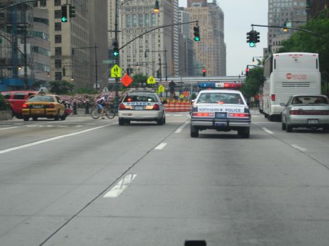 1991 Ford LTD Crown Victoria - North Haven Police sedan and another police car drive down a broad city avenue with a yellow taxi, bus and traffic signals.