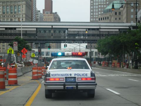 1991 Ford LTD Crown Victoria - white and blue North Haven Police sedan with lightbar drives past orange construction barrels on an urban street lined with high-rises.