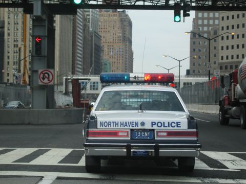 1991 Ford LTD Crown Victoria - North Haven Police sedan with roof lightbar waits at a crosswalk on a downtown street near a red cement mixer truck.