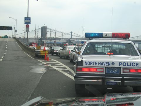 1991 Ford LTD Crown Victoria - North Haven Police sedan leads a line of cars up a bridge ramp toward a suspension bridge under an overcast sky.